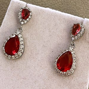 May include: A pair of silver-toned drop earrings featuring a pear-shaped red gemstone at the top and a larger matching pear-shaped red gemstone at the bottom, each surrounded by a halo of small clear white stones set in a metallic frame.
