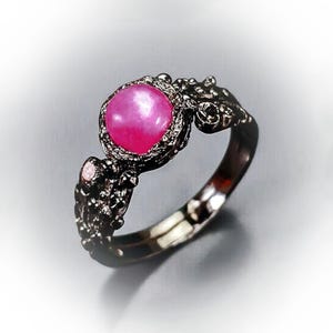 May include: A black ring with a pink gemstone set in a detailed, ornate design. The ring is made of metal and has a polished finish.