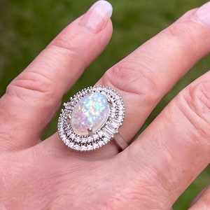 May include: A silver ring featuring an oval, iridescent opal gemstone, surrounded by a halo of small, round, and baguette-cut cubic zirconia stones. The ring is set on a silver band, showcasing a classic and elegant design.