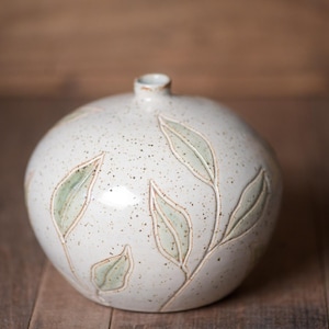 May include: A round ceramic vase with a speckled off-white glaze featuring carved, light green leaf motifs. The vessel has a narrow, circular opening at the top and a smooth, bulbous body that tapers slightly toward the base, showcasing a textured finish.