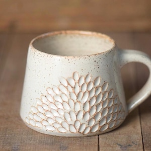 May include: Cream-colored ceramic mug with a textured floral design. The mug has a rounded shape, a handle, and a speckled finish. The floral pattern is carved into the surface, adding a tactile element.