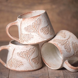 May include: Three ceramic mugs with a speckled off-white glaze and a tapered shape. Each mug features an embossed ginkgo leaf pattern with reddish-brown detailing. The mugs have a sturdy handle and a smooth interior finish, stacked together.
