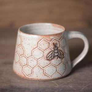 Peut inclure: Une tasse en céramique avec un émail blanc cassé moucheté, présentant un motif en nid d'abeille en relief et une abeille. La tasse possède une base large, un corps effilé et une anse incurvée, avec des tons brun-rouge sur le bord.