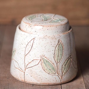 May include: A ceramic jar with a lid, featuring a speckled off-white glaze and hand-carved botanical leaf patterns. The leaves are filled with a soft sage green color, and the jar has a tapered cylindrical shape with a flat top lid.