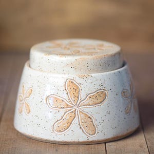 Op de afbeelding: Een keramische botervloot met deksel. De schaal is gebroken wit met een gespikkelde afwerking en heeft een bloemmotief in een lichtbruine kleur. Het deksel heeft ook een bloemmotief.