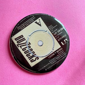 May include: A black and white 45 RPM vinyl record with the band name "Buzzcocks" and the song title "I Don't Mind" printed on the label. The record is from the album "Another Music In A Different Kitchen" released by United Artists Records.