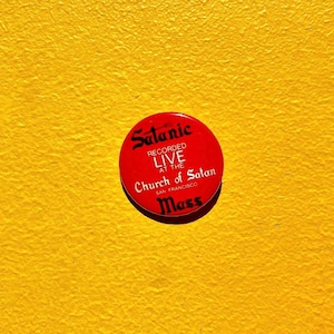 May include: A red button with the word "Satanic" in a stylized font. The button also includes the text "Recorded Live at the Church of Satan San Francisco Mass" in white lettering. The button is set against a textured yellow background.