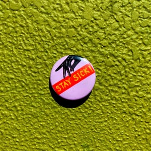 May include: A round, light purple button with the words "STAY SICK!" in red on a diagonal stripe. The button has a black and white graphic above the text, set against a textured, olive-green background.