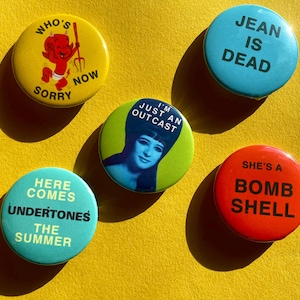 Può includere: Una collezione di cinque spille colorate con vari disegni e testo. Le spille sono gialle, blu, verdi e rosse. Il testo include "Who's Sorry Now", "Jean is Dead" e "She's a Bombshell".