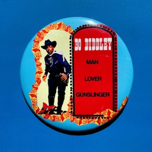 May include: A circular badge with a light blue backdrop. It displays an image of Bo Diddley in a cowboy hat and a red rectangle with the words "BO DIDDLEY, MAN, LOVER, GUNSLINGER". The image is framed by a yellow and red floral design.