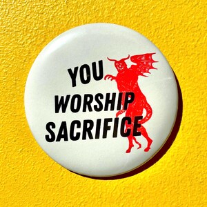May include: A circular white pinback button featuring the black text YOU WORSHIP SACRIFICE and a red graphic of a winged, horned creature. The design is centered on the button, which has a smooth, glossy finish and a standard metal backing.