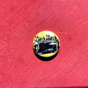 Op de afbeelding: Een gele knoop met de tekst "LOST WEEKEND" en een zwarte auto-afbeelding. De knoop is omgeven door een vlammenontwerp en ligt op een rode, getextureerde ondergrond.