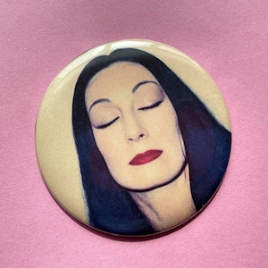 Op de afbeelding: Een ronde knoop met een portret van een vrouw met gesloten ogen, donker haar en rode lippenstift. De knoop heeft een crèmekleurige achtergrond en is geplaatst tegen een roze achtergrond.