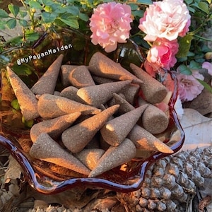 May include: A bowl filled with brown incense cones, surrounded by pine cones and pink roses. The bowl is made of glass with a reddish-brown tint. The text "@MyPaganMaiden" is visible in the image.