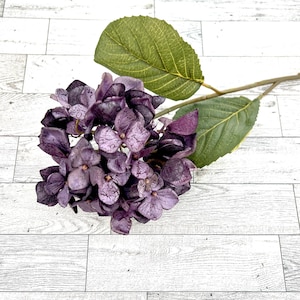 May include: A single stem of artificial purple hydrangea with green leaves. The hydrangea is made of fabric and has a realistic appearance.