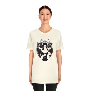 Puede incluir: Una camiseta blanca con una ilustración en blanco y negro de una mujer con una corona y dos cabezas de lobo. La mujer sostiene un cuchillo y tiene una expresión seria en su rostro.