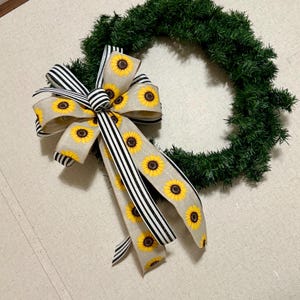 May include: A circular green artificial pine wreath adorned with a large decorative bow. The bow is crafted from beige fabric featuring a yellow sunflower pattern and is accented with black and white vertical striped ribbon streamers.