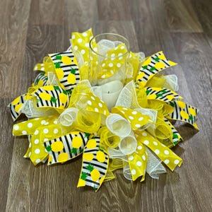 May include: A yellow and white pineapple themed centerpiece with a glass votive holder and a white candle. The centerpiece is made with mesh, ribbon, and polka dot fabric.