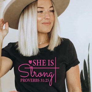 May include: Black t-shirt with a pink graphic that says "She is Strong" with a heart and a cross. The text "Proverbs 31:25" is also included in pink.