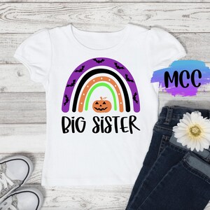 May include: White t-shirt with a purple, black, and green rainbow with bats and a pumpkin. The text "Big Sister" is below the rainbow.