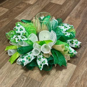 May include: A St. Patrick's Day centerpiece featuring a clear glass candle holder surrounded by green, white, and gold mesh ribbons. The ribbons are adorned with shamrock patterns and glitter accents, with a white candle inside.