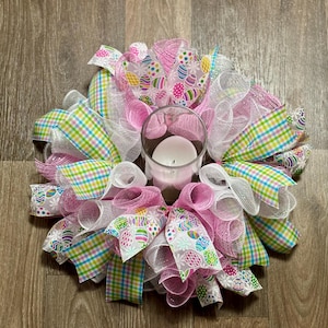 May include: A pink and white Easter wreath with a clear glass candle holder in the center. The wreath is decorated with pink and white mesh, green and white plaid ribbon, and white ribbon with a pastel egg pattern.