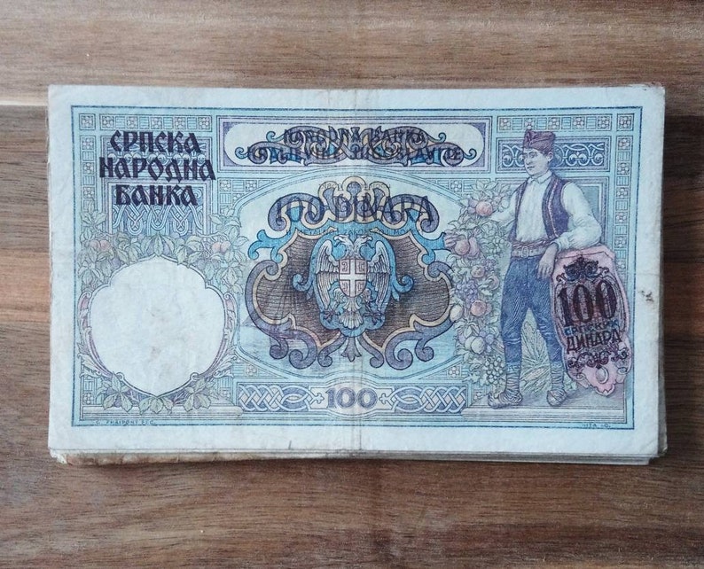 May include: A vintage 100 dinar banknote from the Serbian National Bank. The note features a blue and brown design with a coat of arms and a portrait of a man in traditional clothing.