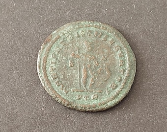 May include: A circular, weathered bronze-toned coin featuring a central embossed figure standing upright, surrounded by a border of worn, illegible text. The surface displays a distinct greenish patina and a textured, aged appearance throughout.