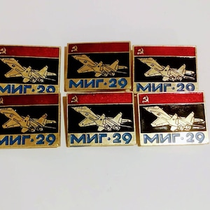 May include: Six gold-colored metal pins with red and black enamel. Each pin features a stylized image of a MiG-29 fighter jet with the text "МИГ-29" in Cyrillic script.