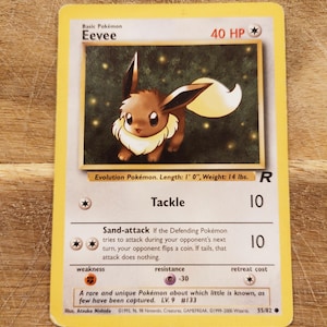 Op de afbeelding: Een Pokémon-verzamelkaart met Eevee, een bruin en crèmekleurig wezen met grote oren en een pluizige staart. De kaart toont de tekst "Eevee", "Tackle" en "Sand-attack", samen met spelgerelateerde informatie. De kaart is 30,48 cm lang en weegt 6,35 kg.
