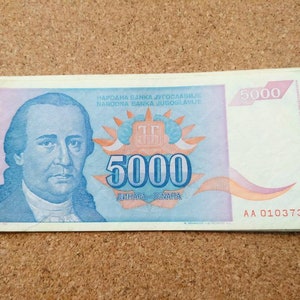 May include: A 5,000 dinar note from the National Bank of Yugoslavia. The note features a portrait of a man wearing a white collar and a blue jacket. The note is in good condition and has a serial number of AA 0103733.