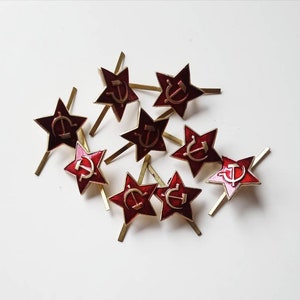 Puede incluir: Un grupo de diez pines de estrella roja con un diseño de martillo y hoz dorado. Los pines están dispersos sobre una superficie blanca.
