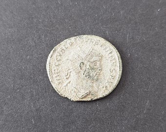 May include: A circular silver-toned ancient coin featuring a profile portrait of a bearded figure wearing a radiate crown. The coin has a textured, weathered surface with visible embossed lettering along the outer edge surrounding the central bust.