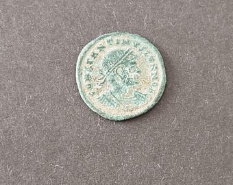 May include: A circular, weathered bronze-toned coin featuring a profile portrait of a figure wearing a diadem. The surface displays a green patina with visible raised lettering along the outer edge, depicting an ancient historical artifact.