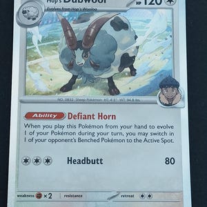 May include: A Hop's Dubwool trading card featuring a grey and white sheep Pokémon with large brown horns. The card displays 120 HP, the Defiant Horn ability, and the Headbutt attack. Text includes card details, stats, and a short flavour description.