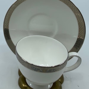May include: A white porcelain teacup and saucer with a gold and silver patterned rim. The saucer has a wide rim with a decorative pattern. The teacup has a small handle and a gold and silver patterned band around the rim.