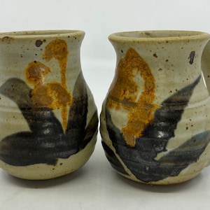 May include: Two handmade ceramic mugs with a beige speckled finish. Each mug features a unique design with brown and black leaf-like patterns and a touch of orange. One mug has a handle.