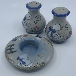 May include: A set of three ceramic salt and pepper shakers and a candle holder, all featuring a white duck design with blue accents. The shakers have a blue rim and the candle holder has a blue center.