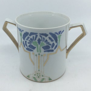 May include: White porcelain cup with gold trim and a blue and green floral design. The cup has two gold handles.