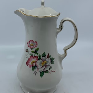 May include: White ceramic creamer with a gold trim and floral design. The creamer has a lid and a handle.