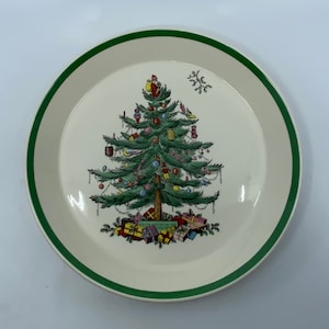 May include: A white ceramic Christmas plate with a green rim. The plate features a colorful illustration of a Christmas tree decorated with ornaments, lights, and presents.