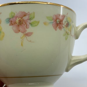 May include: A white teacup with a gold rim and a floral pattern featuring pink, yellow, and green flowers. The cup has a handle and a delicate design.