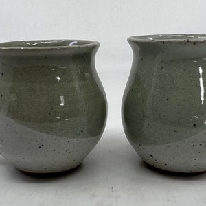 May include: Two handmade ceramic mugs. Each mug has a rounded body with a handle and a two-tone color scheme: a light gray bottom and a darker gray top. The mugs have a speckled texture.