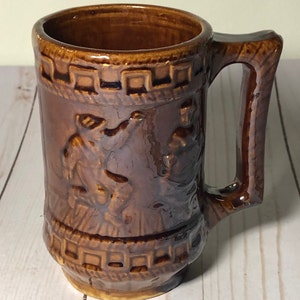 May include: A brown ceramic mug with a raised design of two figures. The mug has a handle and a brown glaze.