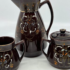May include: A brown ceramic tea set with gold accents and floral designs. The set includes a teapot, creamer, and sugar bowl.