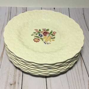 May include: A stack of eight white ceramic dinner plates with a raised embossed pattern and a floral design in the center. The floral design features pink, yellow, blue, and red flowers.