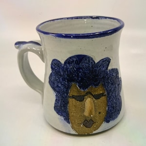 May include: A white ceramic mug with a blue rim and a hand-painted design of a face with blue hair and sunglasses. The face is brown with a brown nose and a black mouth.