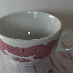 May include: A white ceramic teacup with a pink and white patterned design. The pattern features a geometric design with a floral motif.