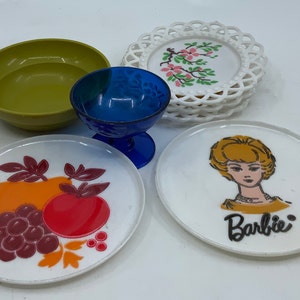 May include: A set of vintage Barbie doll dishes, including a green bowl, a blue glass, a white plate with a fruit design, a white plate with a Barbie design, and a stack of white plates with a floral design.