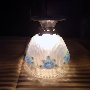 Może przedstawiać: Zabytkowa szklana lampa sufitowa. Lampa ma biały, żebrowany klosz z niebieskimi i zielonymi kwiatowymi akcentami. Oprawa jest podświetlona, ​​rzucając ciepłą poświatę. Podstawa jest z przezroczystego szkła.